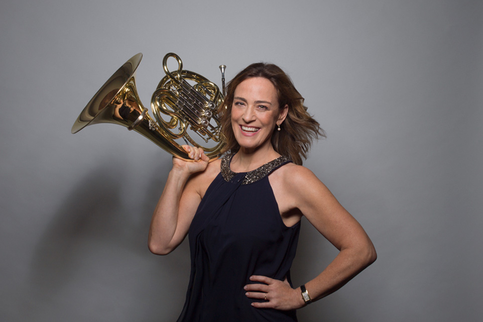 Sarah Willis holding a French horn, wearing a black top, smiling at the camera.